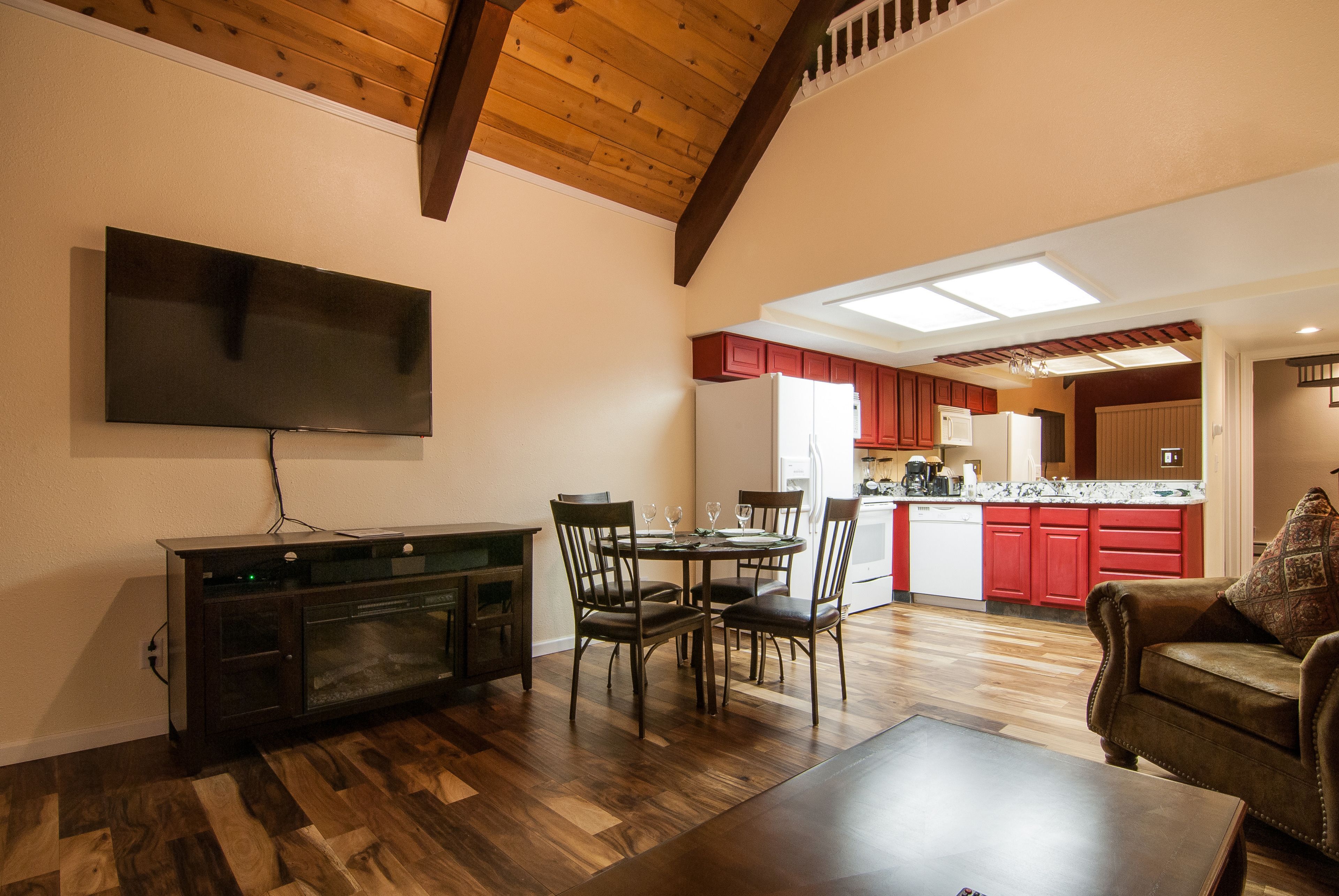 A living room with a TV, dining table, chairs, and a sofa. A kitchen with red cabinets and a white refrigerator is visible in the background.