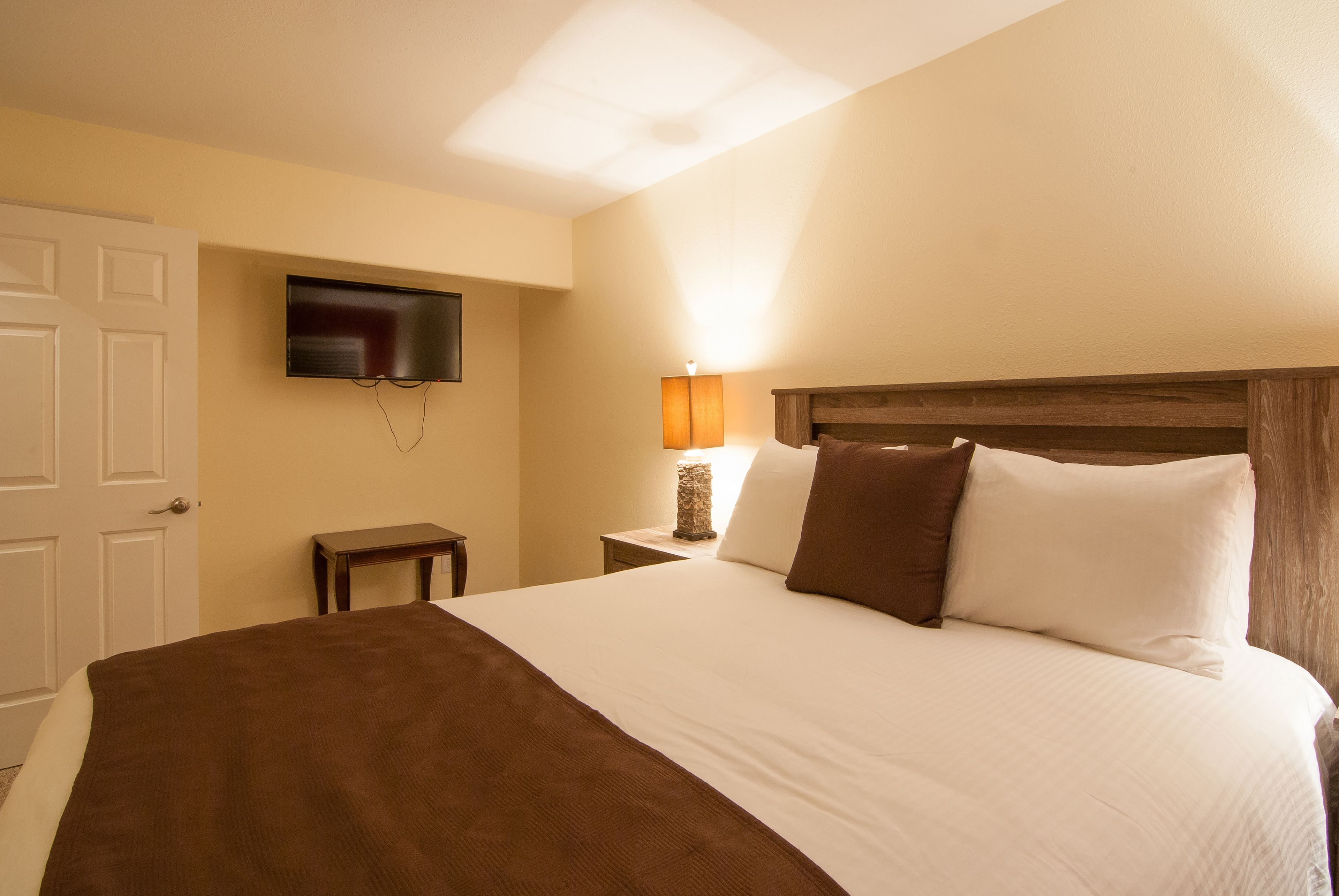 A hotel room with a large bed, a television mounted on the wall, a bedside table with a lamp, and a small nightstand.