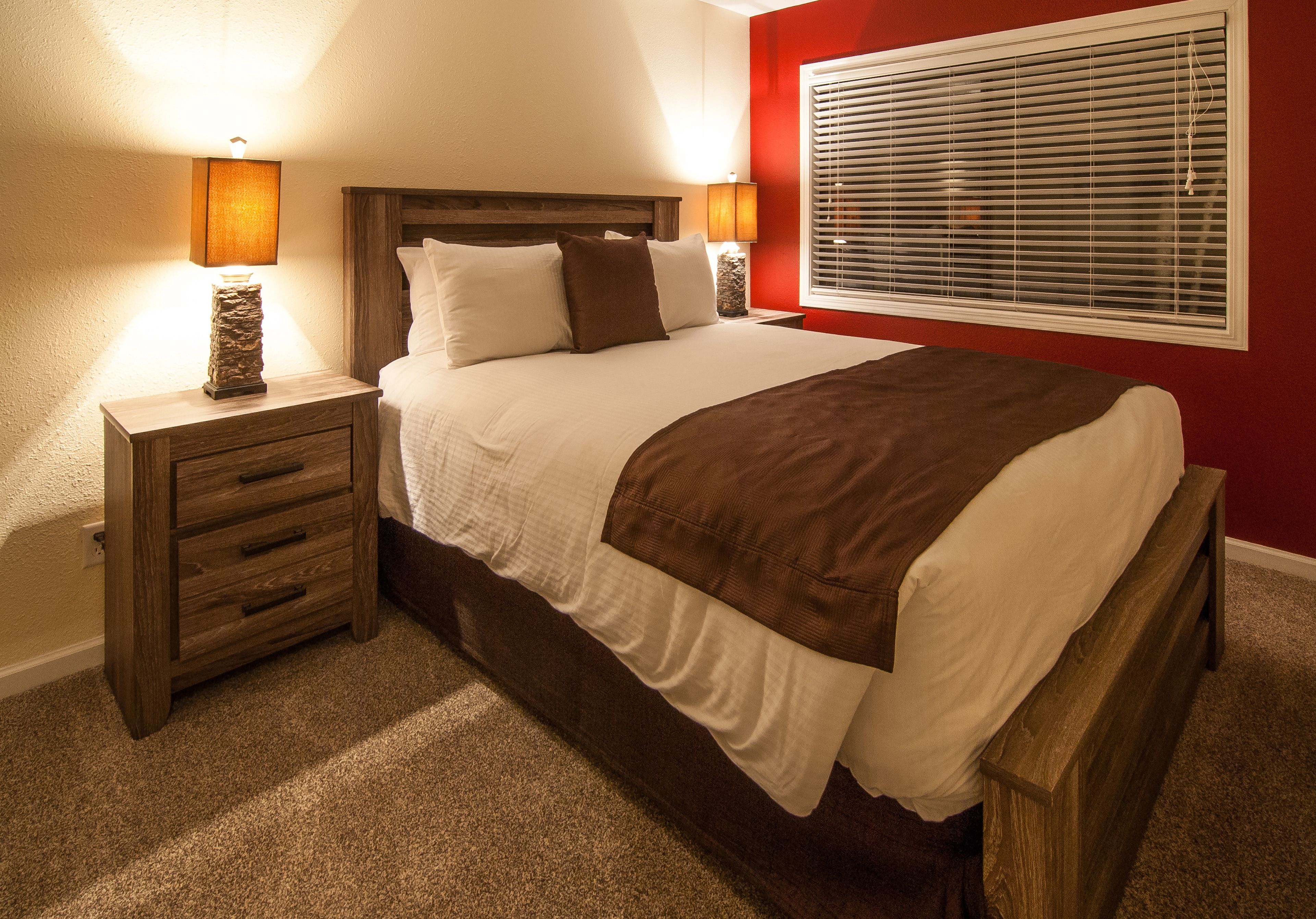 Una cama bien hecha con cabecera de madera, dos mesitas de noche con lámparas y una ventana con persianas.