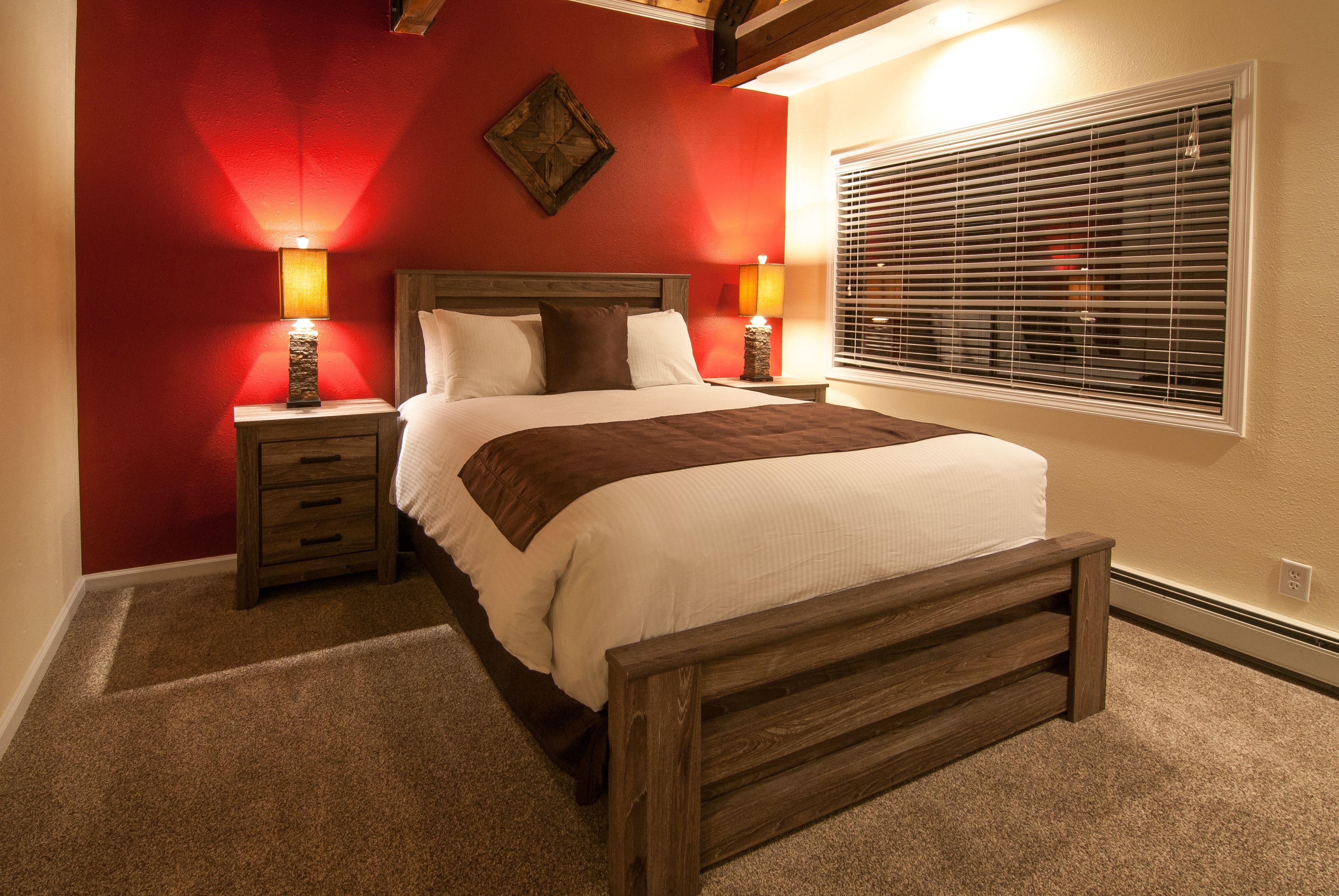 A bedroom with a wooden bed, two bedside tables with lamps, a window with blinds, and a red accent wall.