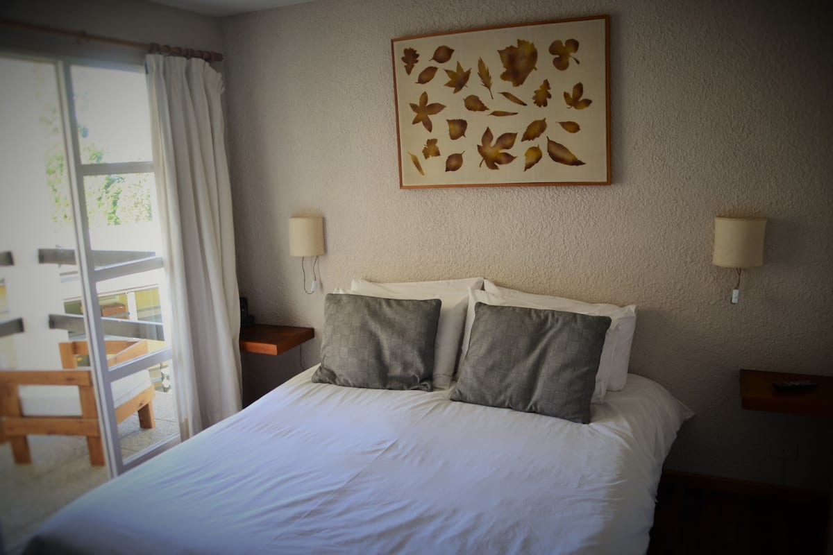 A bedroom with a bed, two bedside lamps, a wall-mounted artwork of leaves, and a view of a balcony with a chair.