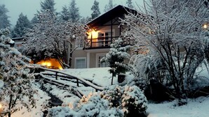A snow-covered landscape with a wooden bridge, a house with a warm light, and snow-laden trees. - Residencia Pioneros (San Carlos de Bariloche)