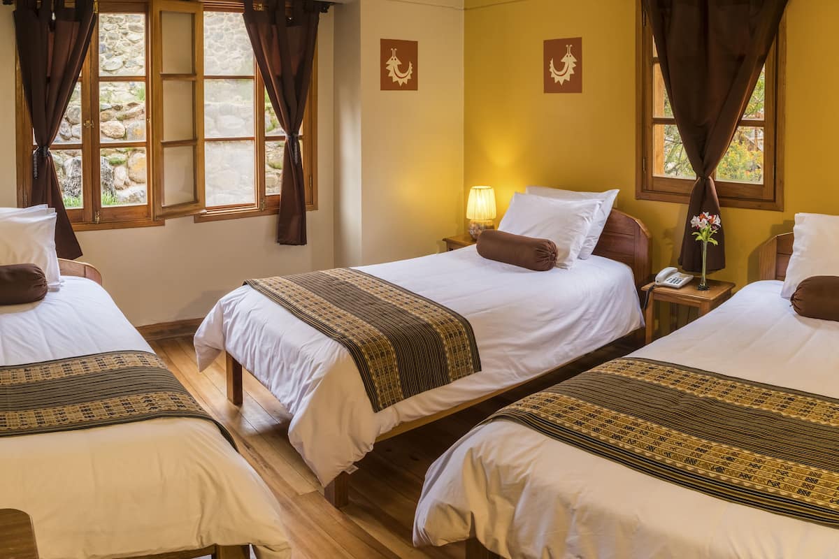 A hotel room with three single beds, each with a brown pillow and a patterned bedspread. There is a nightstand with a lamp and a telephone on one side. The room has a stone wall and a window with curtains.