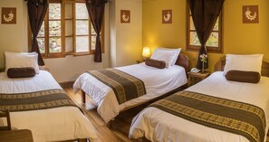 A hotel room with three single beds, each with a brown pillow and a patterned bedspread. There is a nightstand with a lamp and a telephone on one side. The room has a stone wall and a window with curtains. - Hotel Pakaritampu (Ollantaytambo)