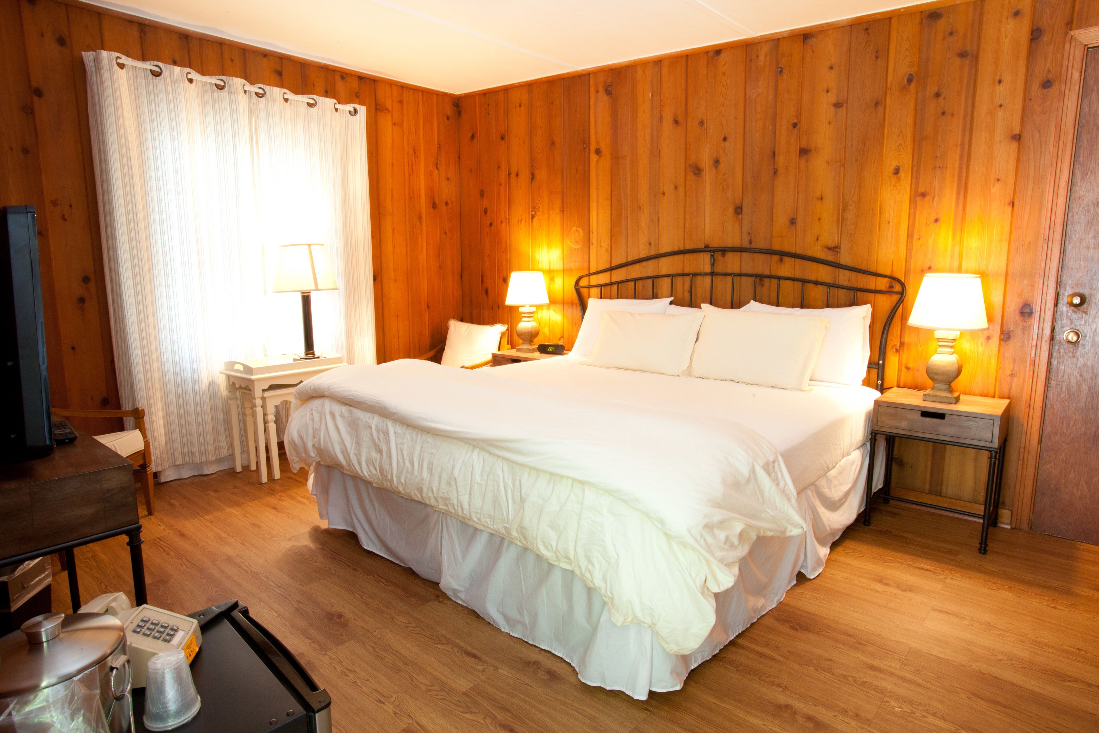 A neatly arranged hotel room with a wooden paneled wall, a large bed with white bedding, two bedside tables with lamps, a small desk with a chair, and a television on a wooden dresser.