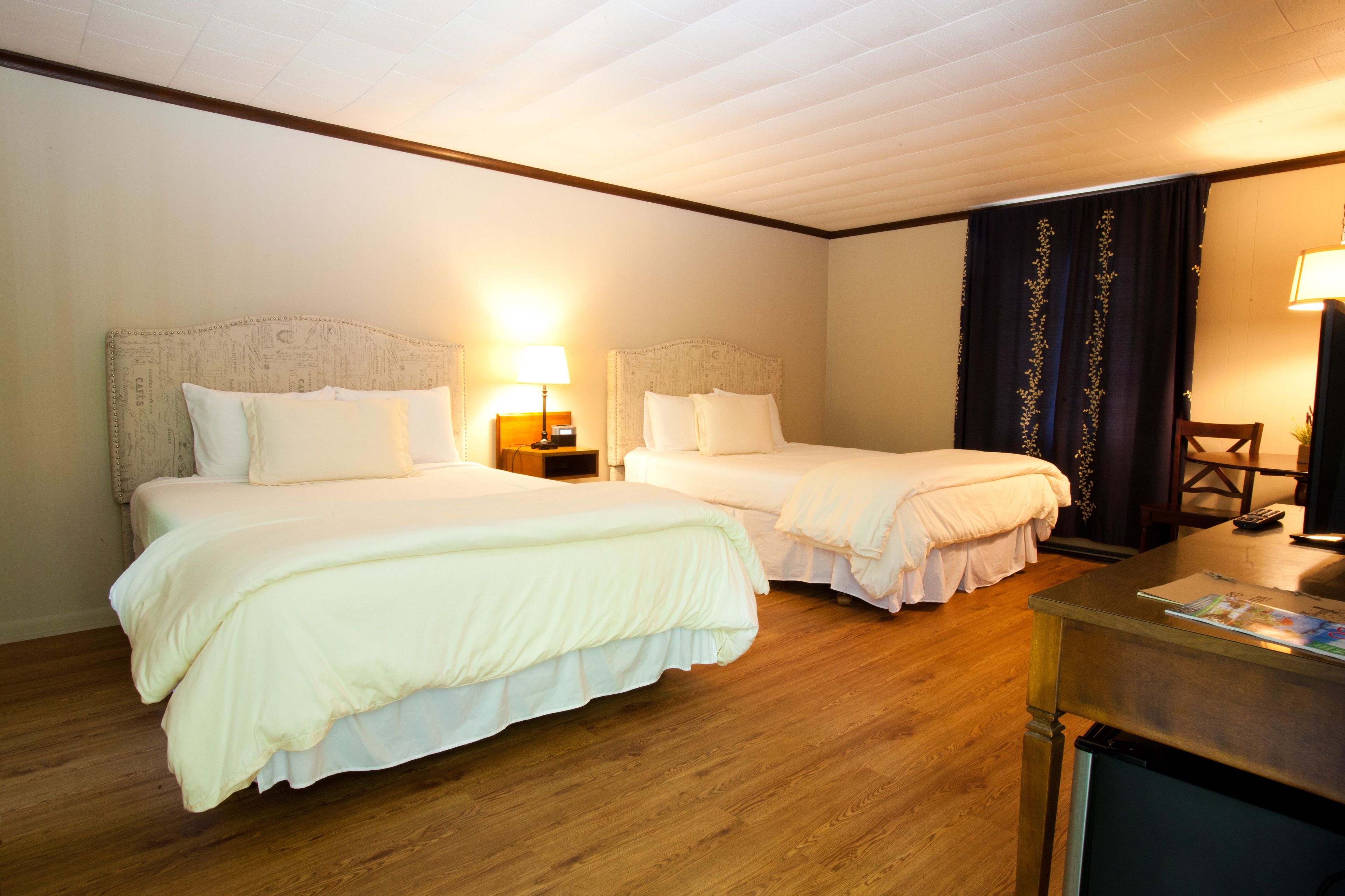 A hotel room with two beds, wooden flooring, a desk with a lamp, and a television.
