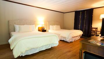 A hotel room with two beds, wooden flooring, a desk with a lamp, and a television.