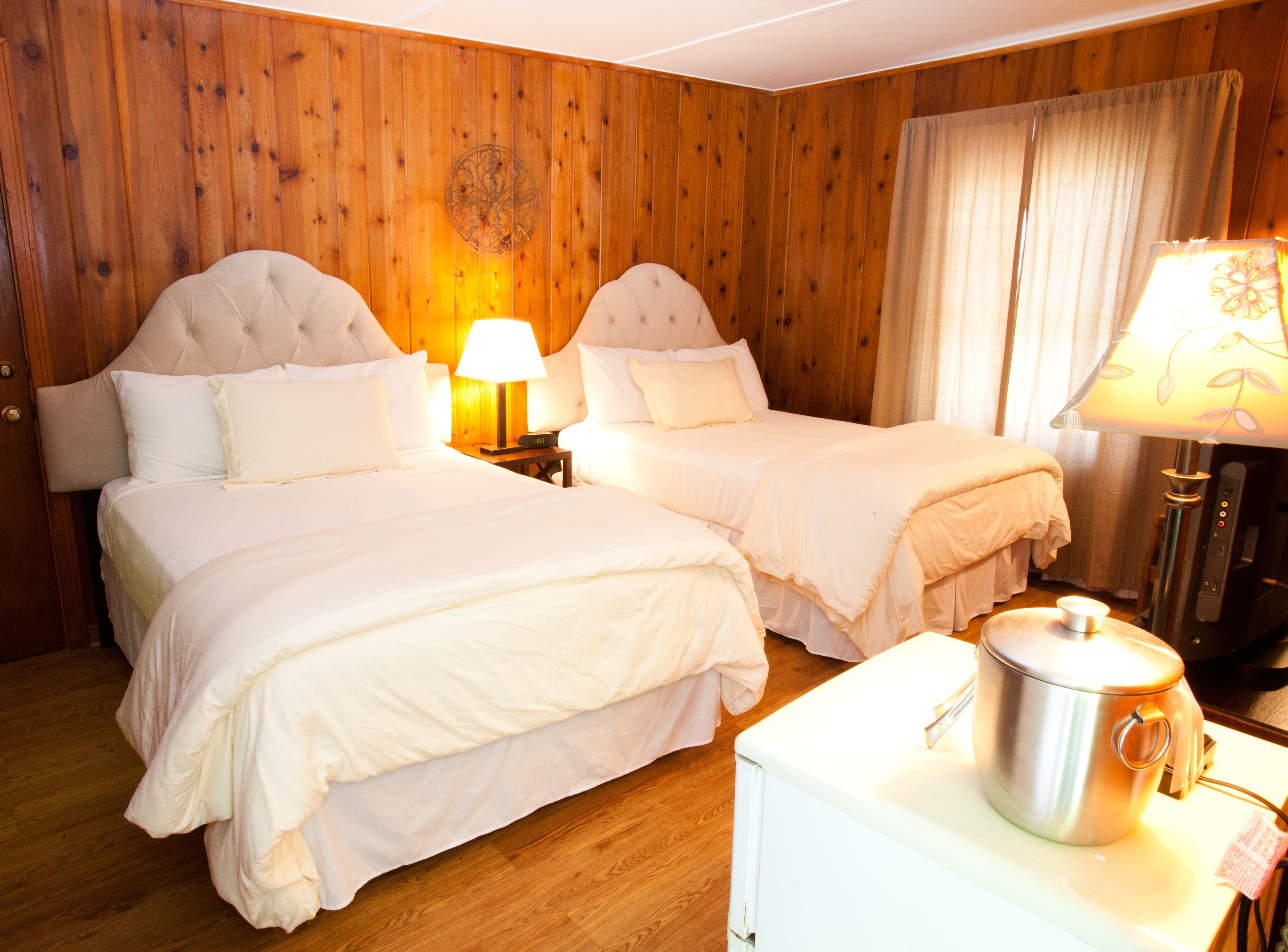 A hotel room with two beds, wooden paneling, a small table with a kettle, and a lamp.