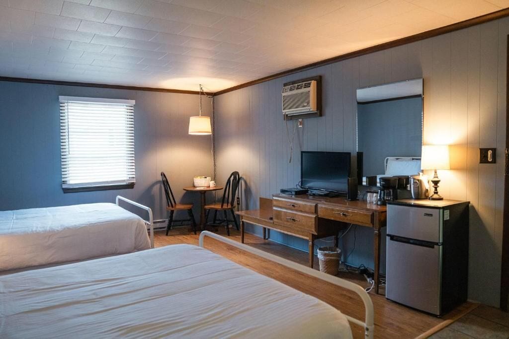 Een hotelkamer met twee bedden, een bureau, een televisie, een koelkast en een raam met jaloezieën.