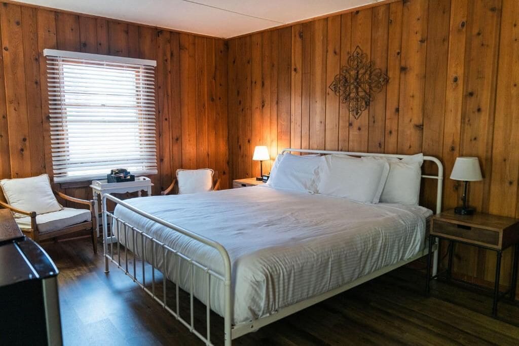 Habitación con una cama individual, pared de paneles de madera, mesita de noche con lámpara, una silla y ventana con persianas.