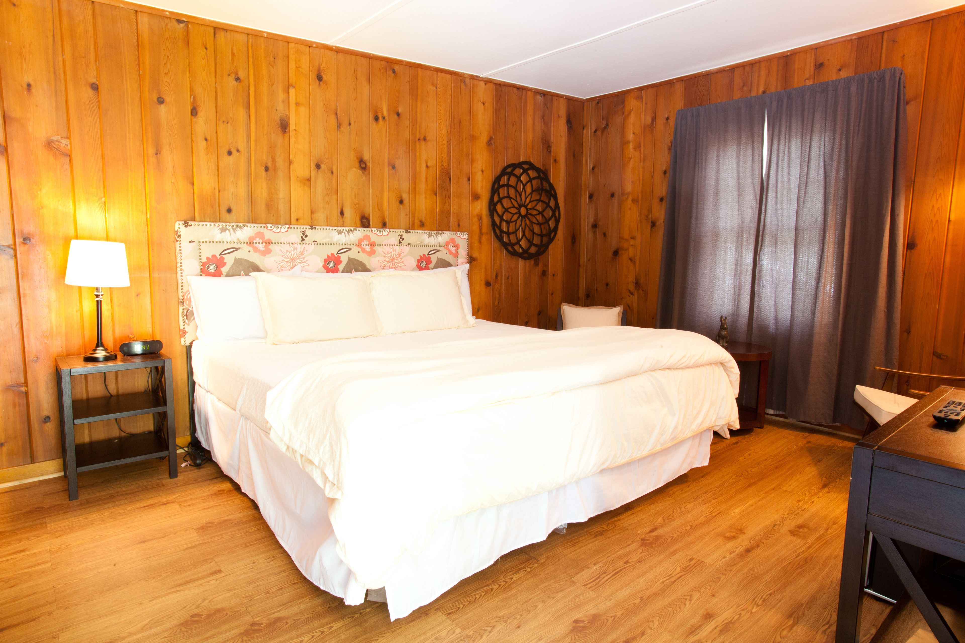 A bedroom with a wooden floor, a bed with white bedding, a nightstand with a lamp, and a wall decoration.