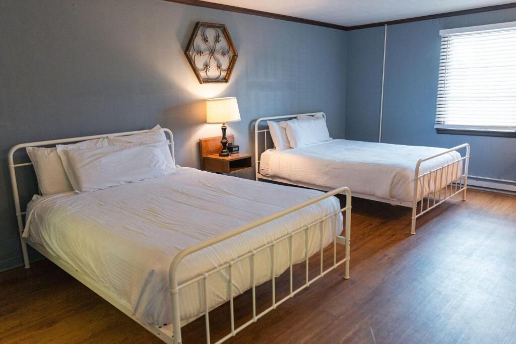 A room with two single beds, a bedside table with a lamp, a wall-mounted artwork, and a window with blinds.
