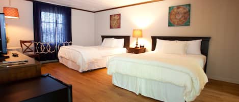 A hotel room with two beds, wooden flooring, a desk with a computer, and a window with curtains.