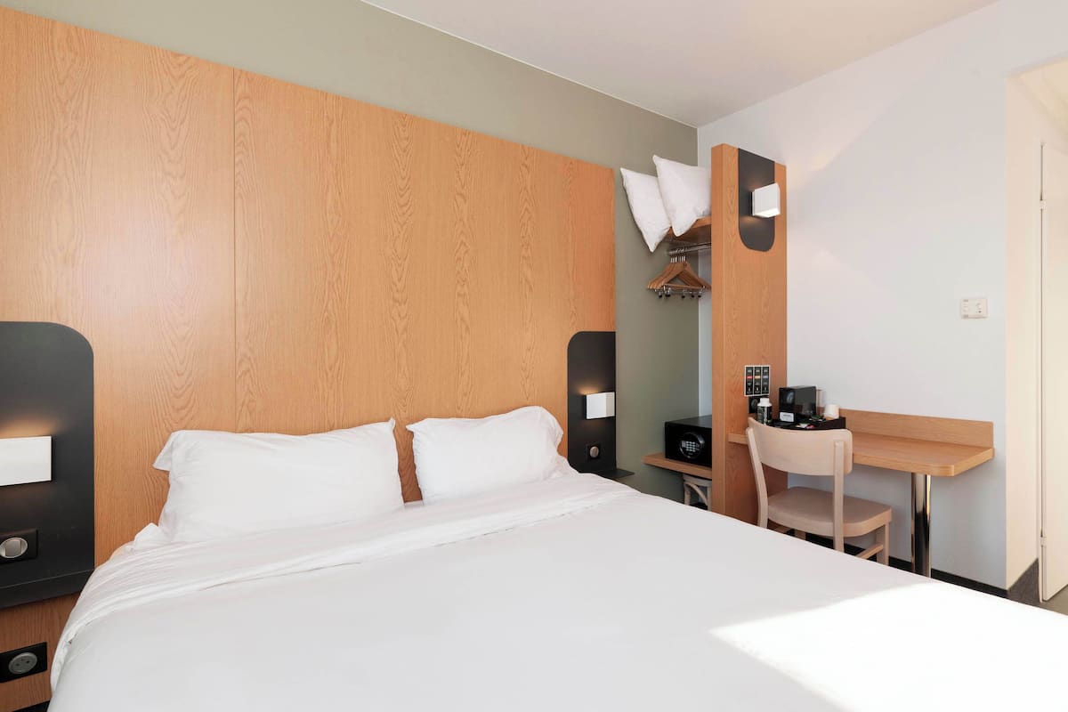 Una habitación de hotel con cabecera de madera, cama con ropa blanca, escritorio con silla y una mesita con cafetera.