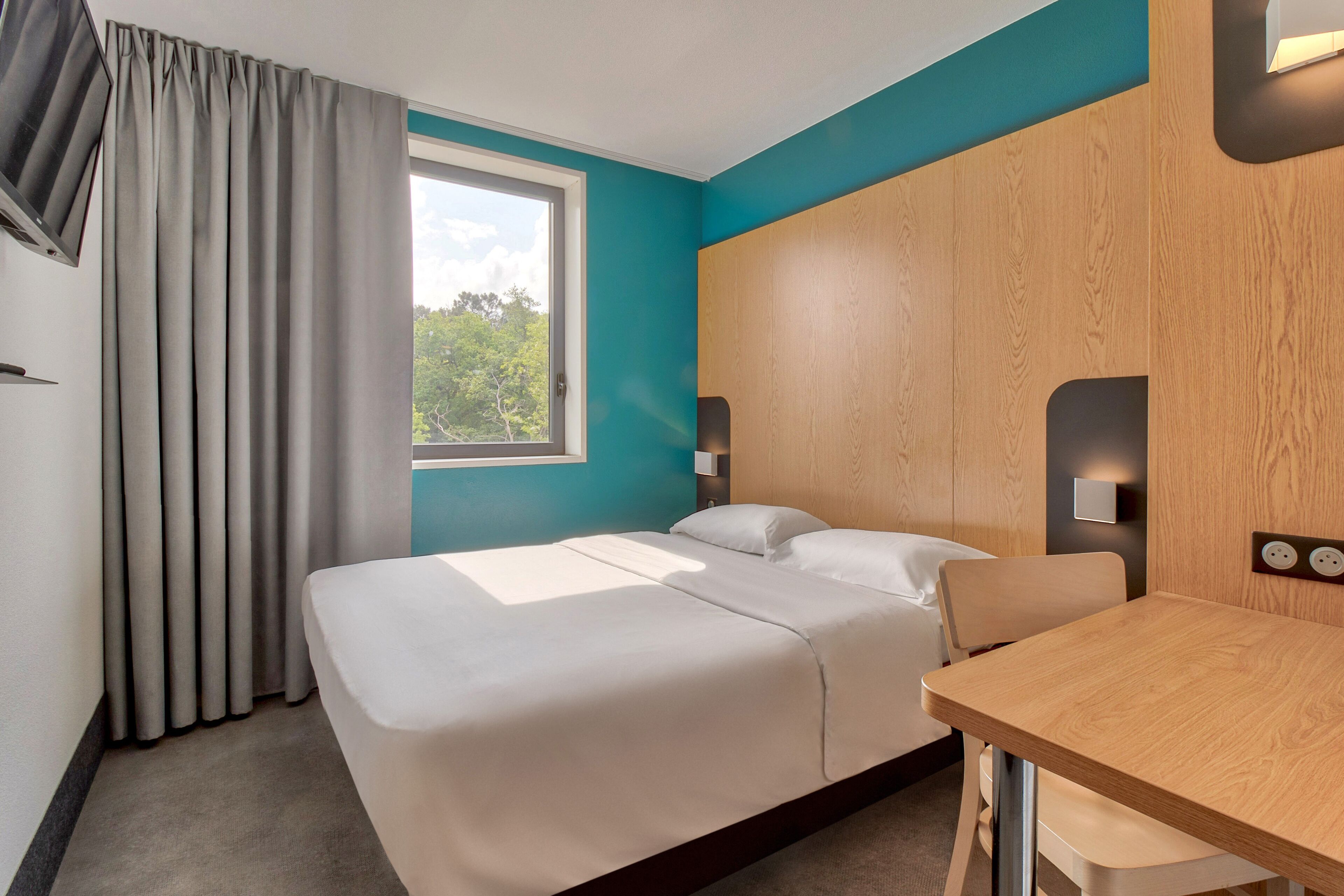 A hotel room with a bed, a table, and a window with curtains.