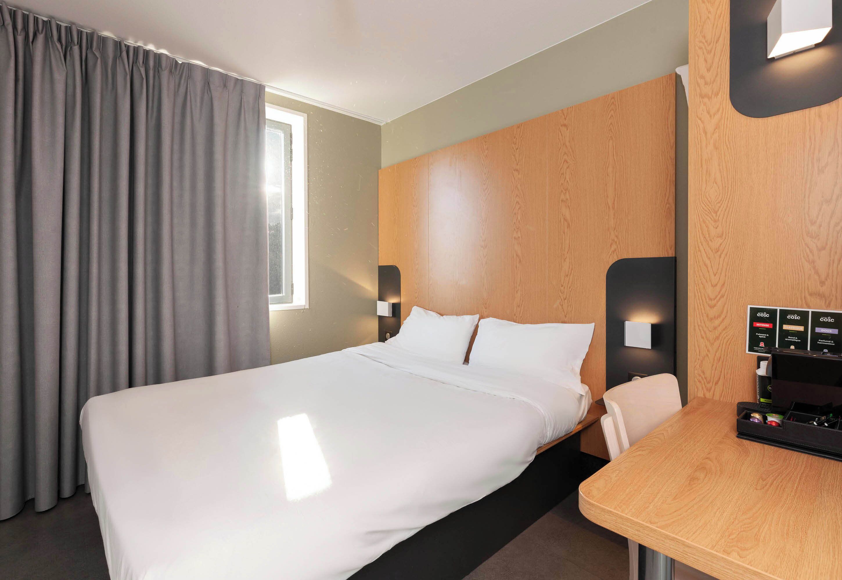 Une chambre d’hôtel avec un grand lit, une tête de lit en bois, une table de chevet avec une machine à café et une fenêtre avec des rideaux.