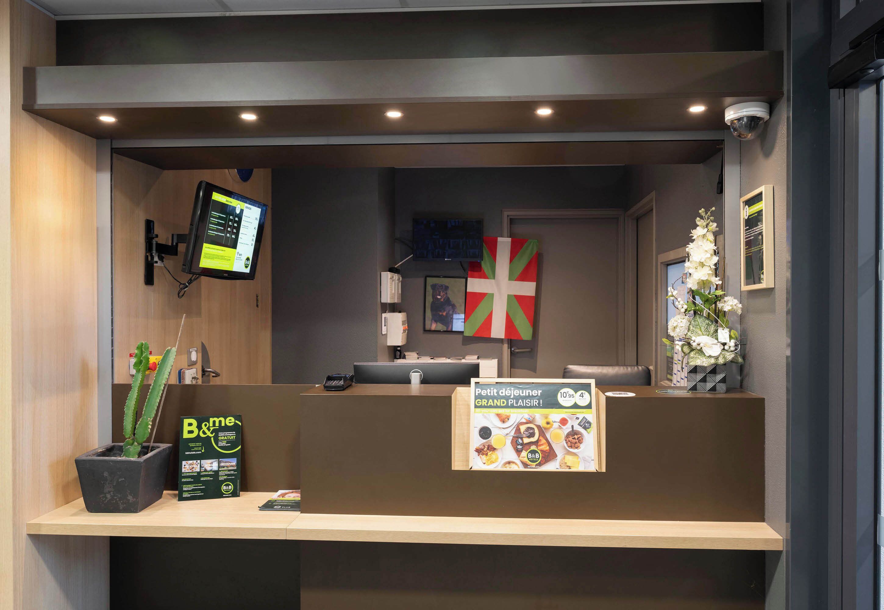 Un comptoir d’accueil doté d’un écran numérique, un drapeau britannique, un panneau indiquant le menu du petit-déjeuner et une plante en pot.