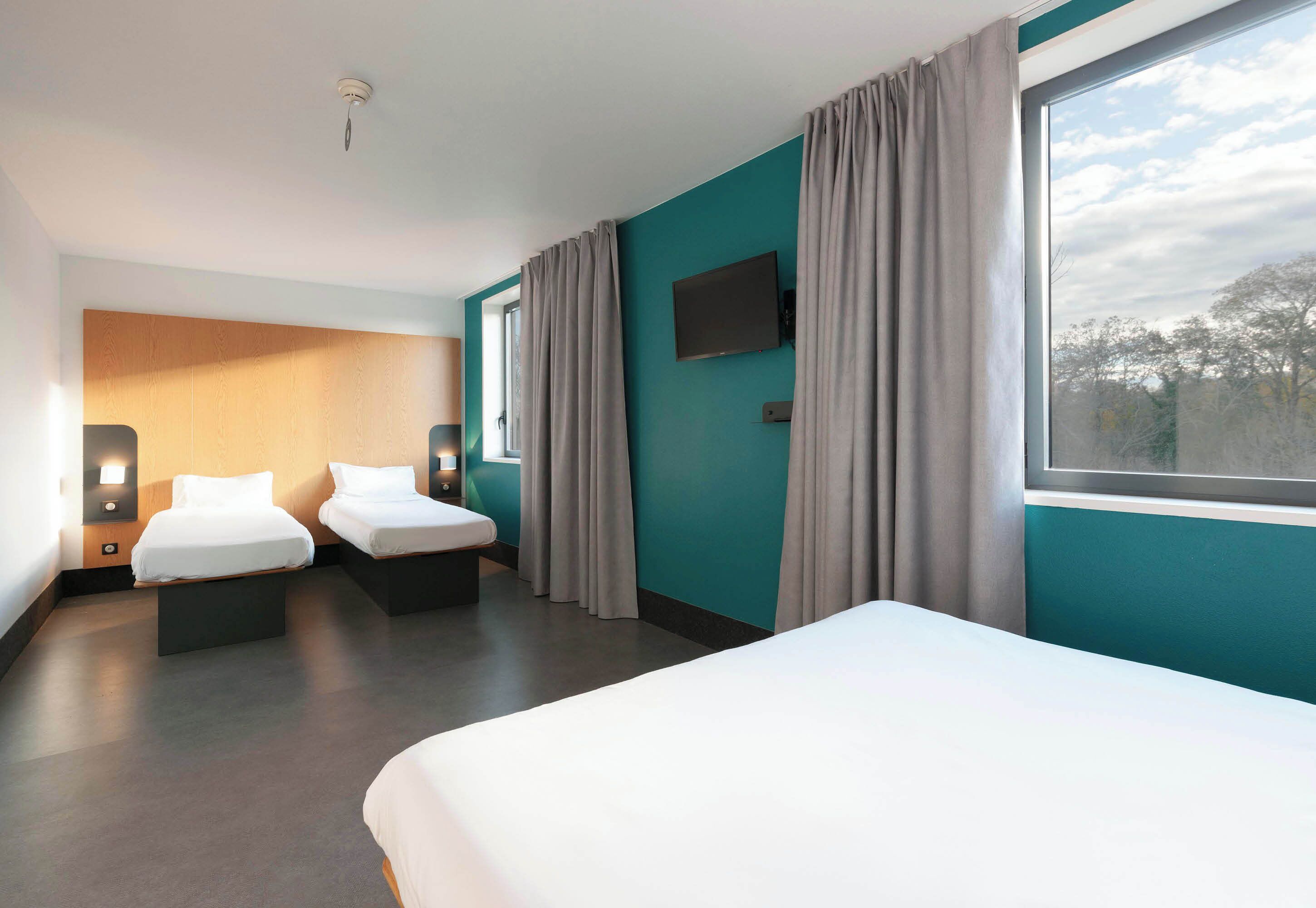 Una habitación de hotel moderna con dos camas individuales, un ventanal con cortinas, un televisor de pantalla plana y un cabecero de madera.