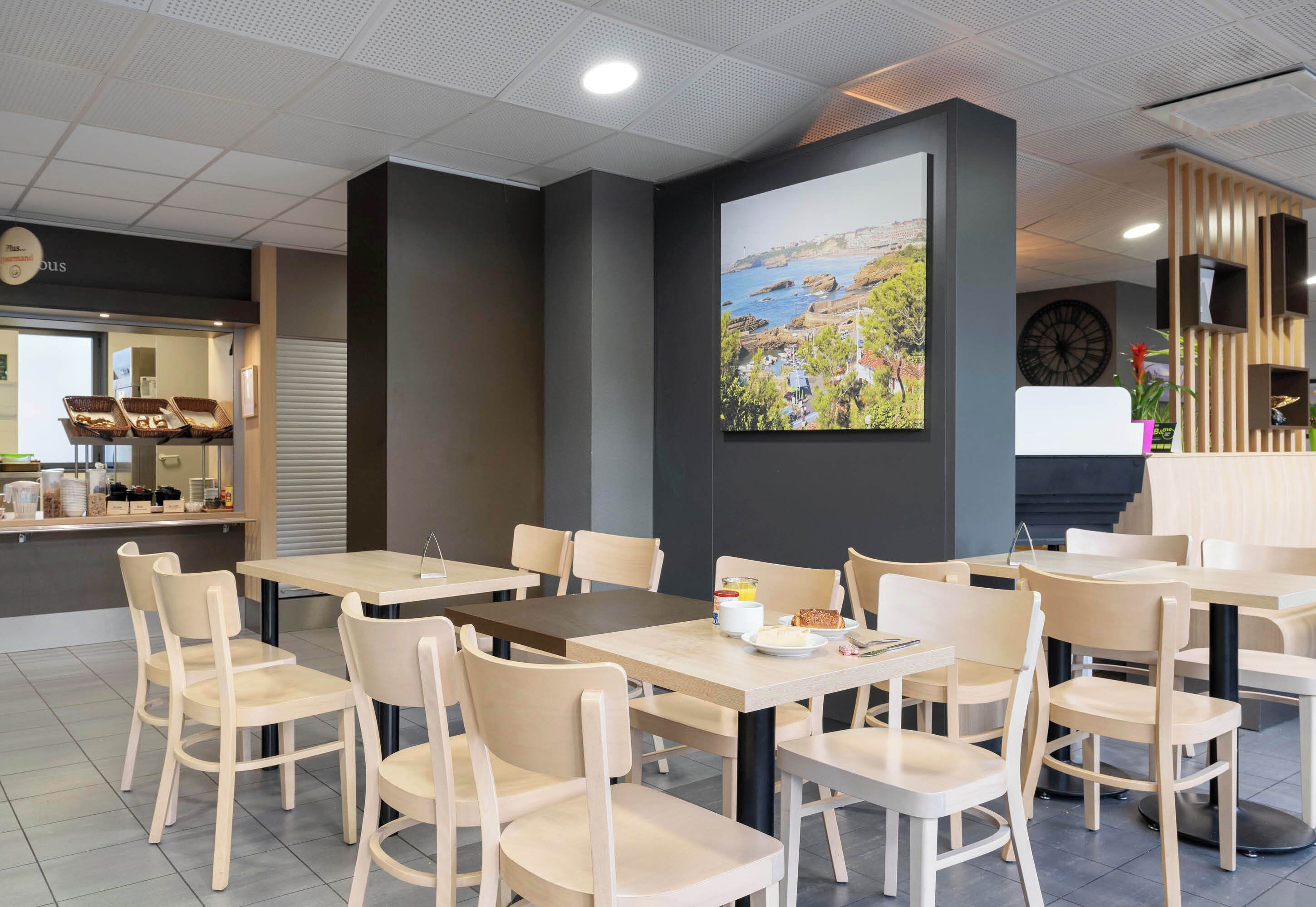 A modern cafe with wooden tables and chairs, a large painting of a coastal landscape, and a counter with baked goods.