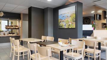 Un café moderno con mesas y sillas de madera, un cuadro grande de un paisaje costero y una barra con productos de panadería.
