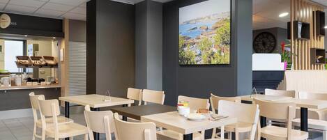 A modern cafe with wooden tables and chairs, a large painting of a coastal landscape, and a counter with baked goods.
