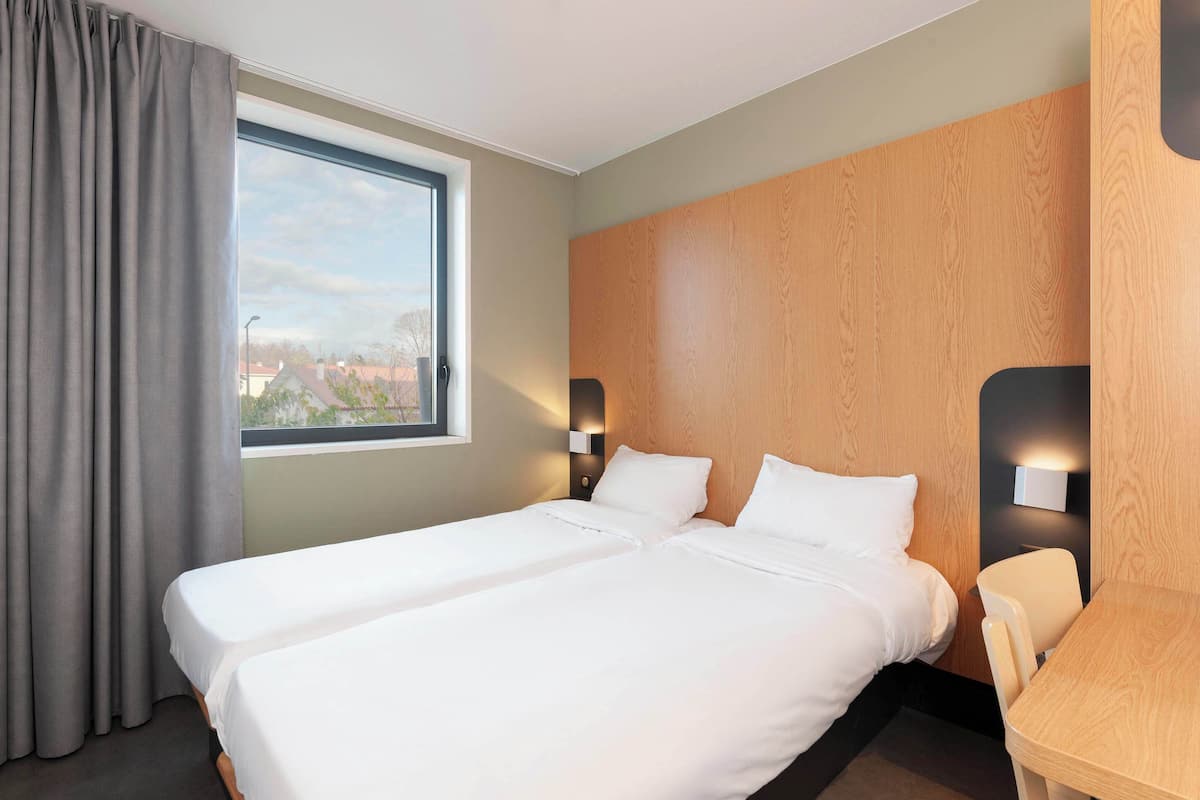 Habitación de hotel con dos camas individuales, un ventanal grande con cortinas, un cabecero de madera, una mesita pequeña y dos lámparas fijadas a la pared.