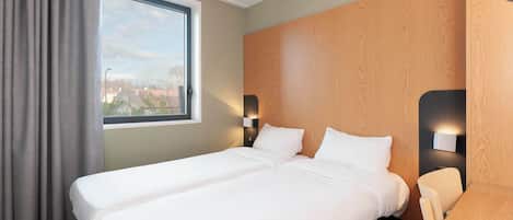 A hotel room with two single beds, a large window with curtains, a wooden headboard, a small table, and two wall-mounted lamps.