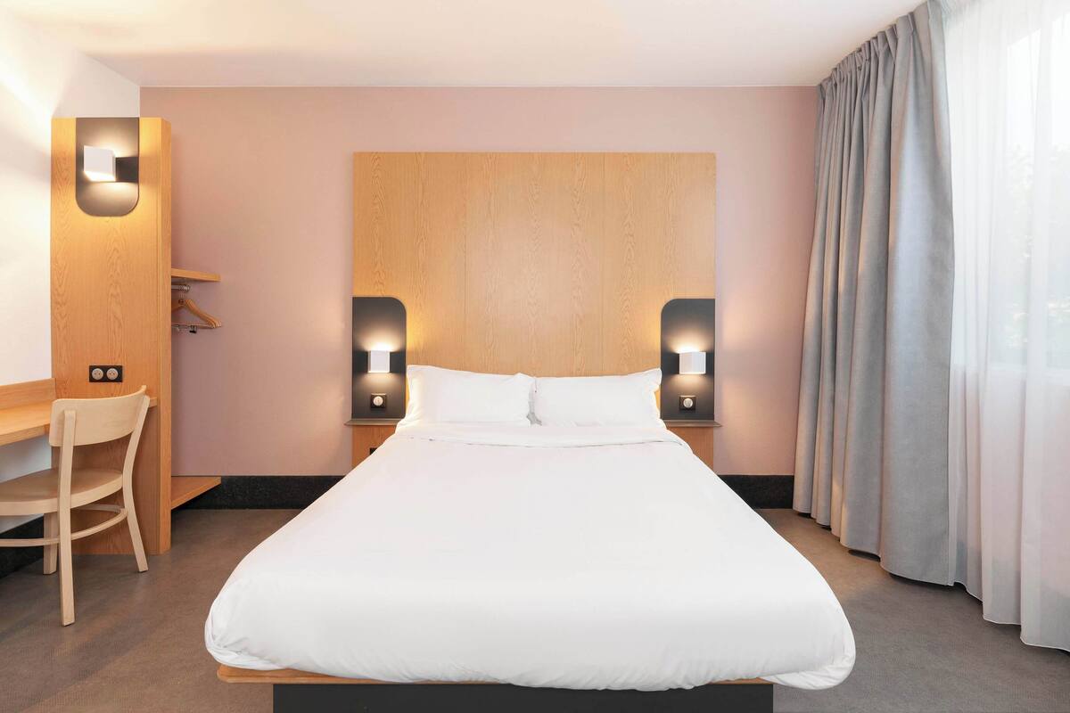 Habitación de hotel con una cama grande, dos lámparas de noche, cabecera de madera, escritorio con silla y ventana con cortinas.