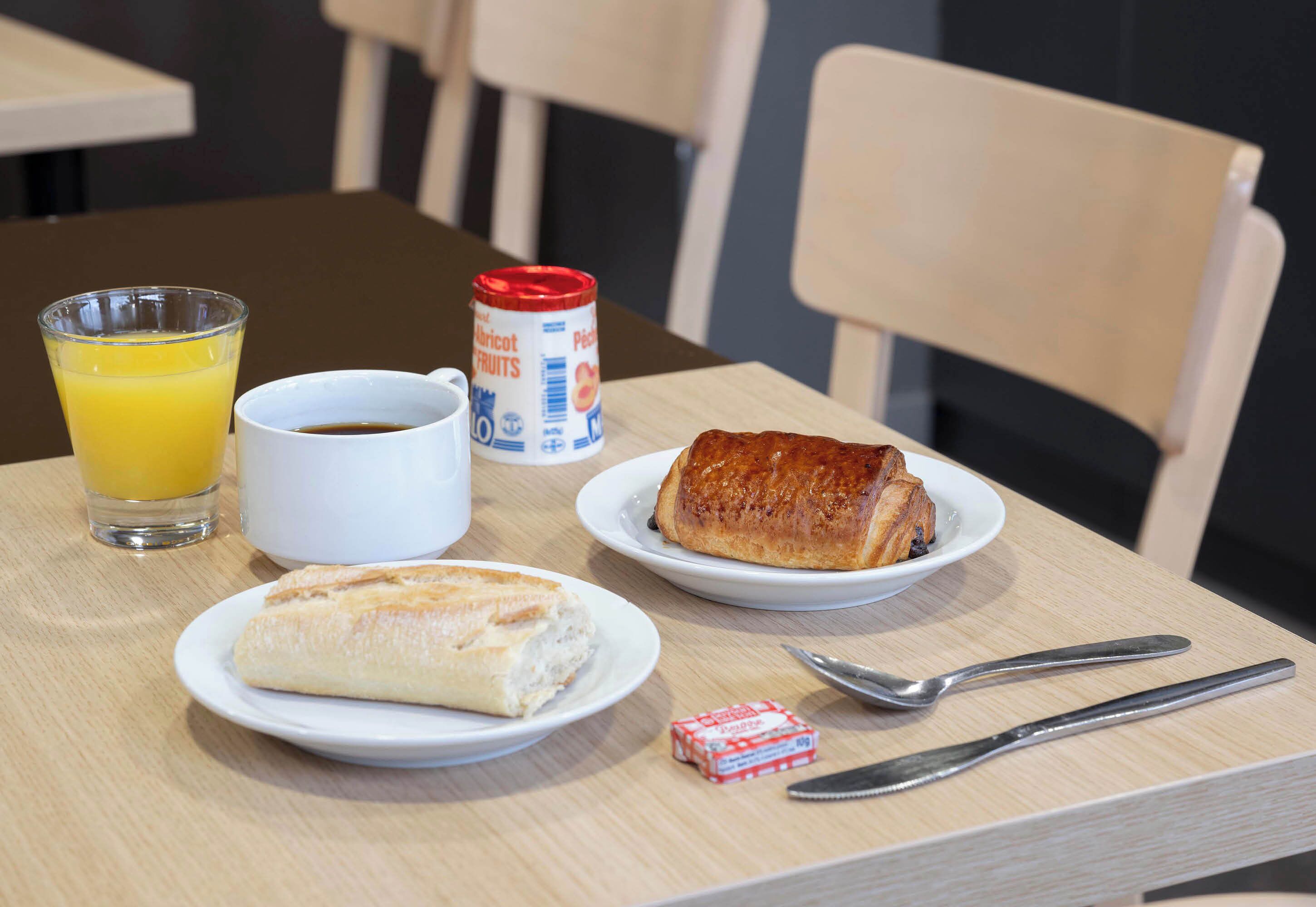 Un desayuno con un croissant, un vaso de zumo de naranja, una taza de café y una lata de leche condensada sobre una mesa.