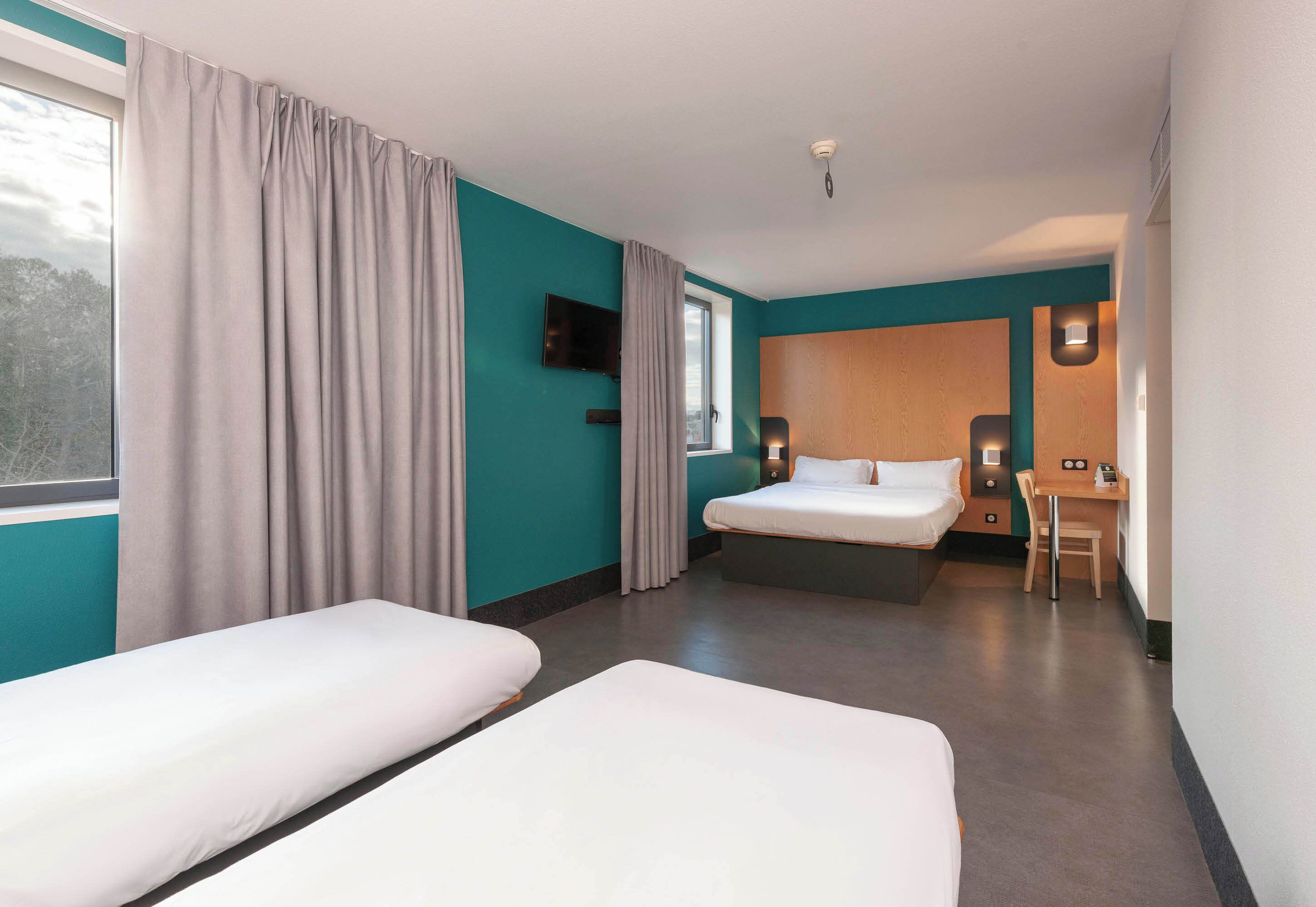 A hotel room with two single beds, a desk, a chair, a TV, and a window with curtains.