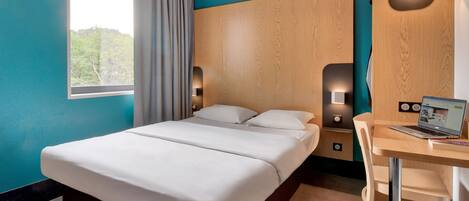 A modern hotel room with a large bed, a desk with a laptop, and a view of the outdoors through a window.