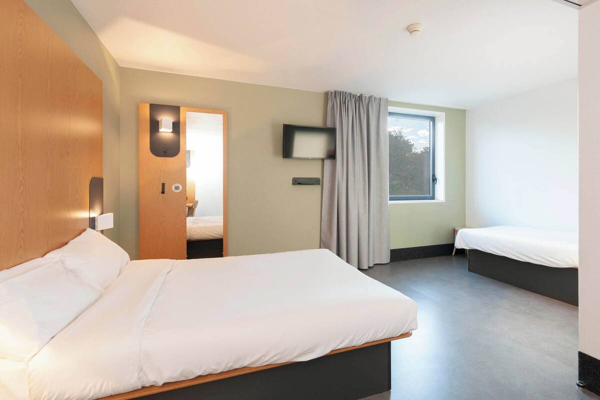 Una habitación de hotel moderna con una cama grande, una cama más pequeña, un televisor y un ventanal con cortinas.