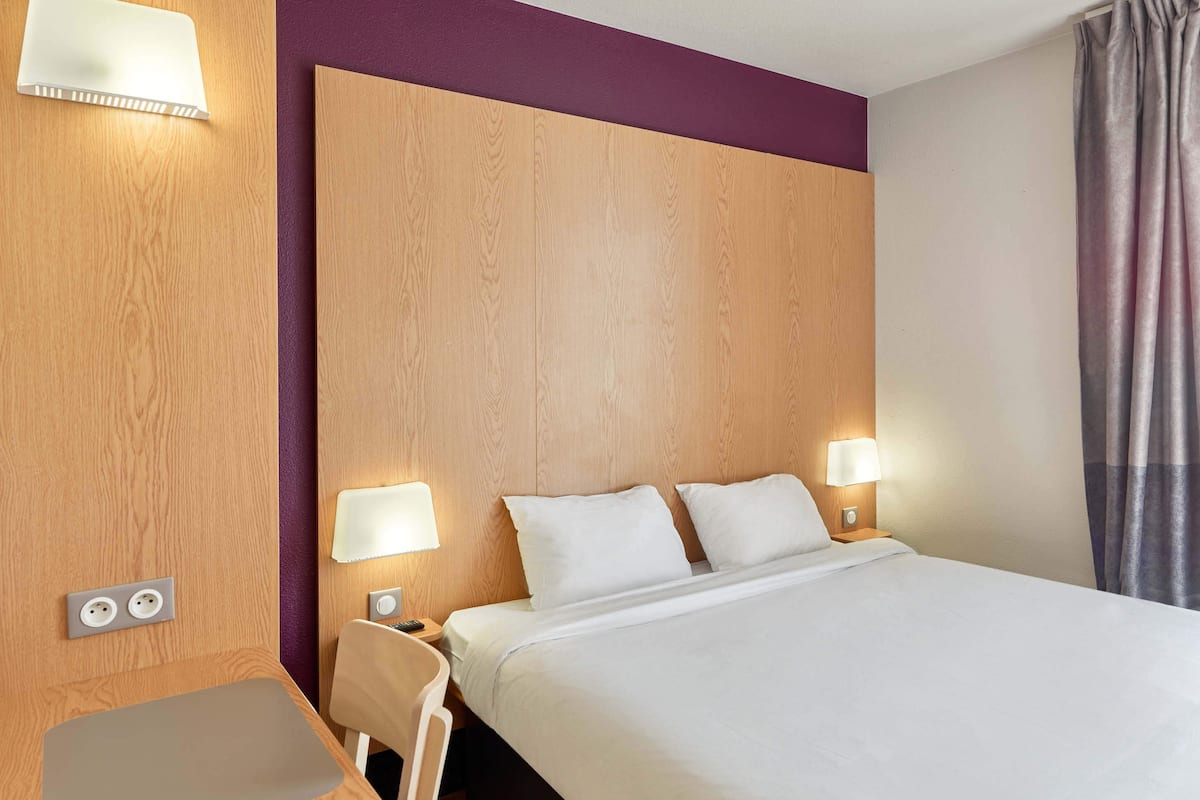 Ein Hotelzimmer mit Holz-Kopfteil, zwei Nachttischlampen, einem Bett mit weißer Bettwäsche, einem kleinen Tisch und einem Sessel.