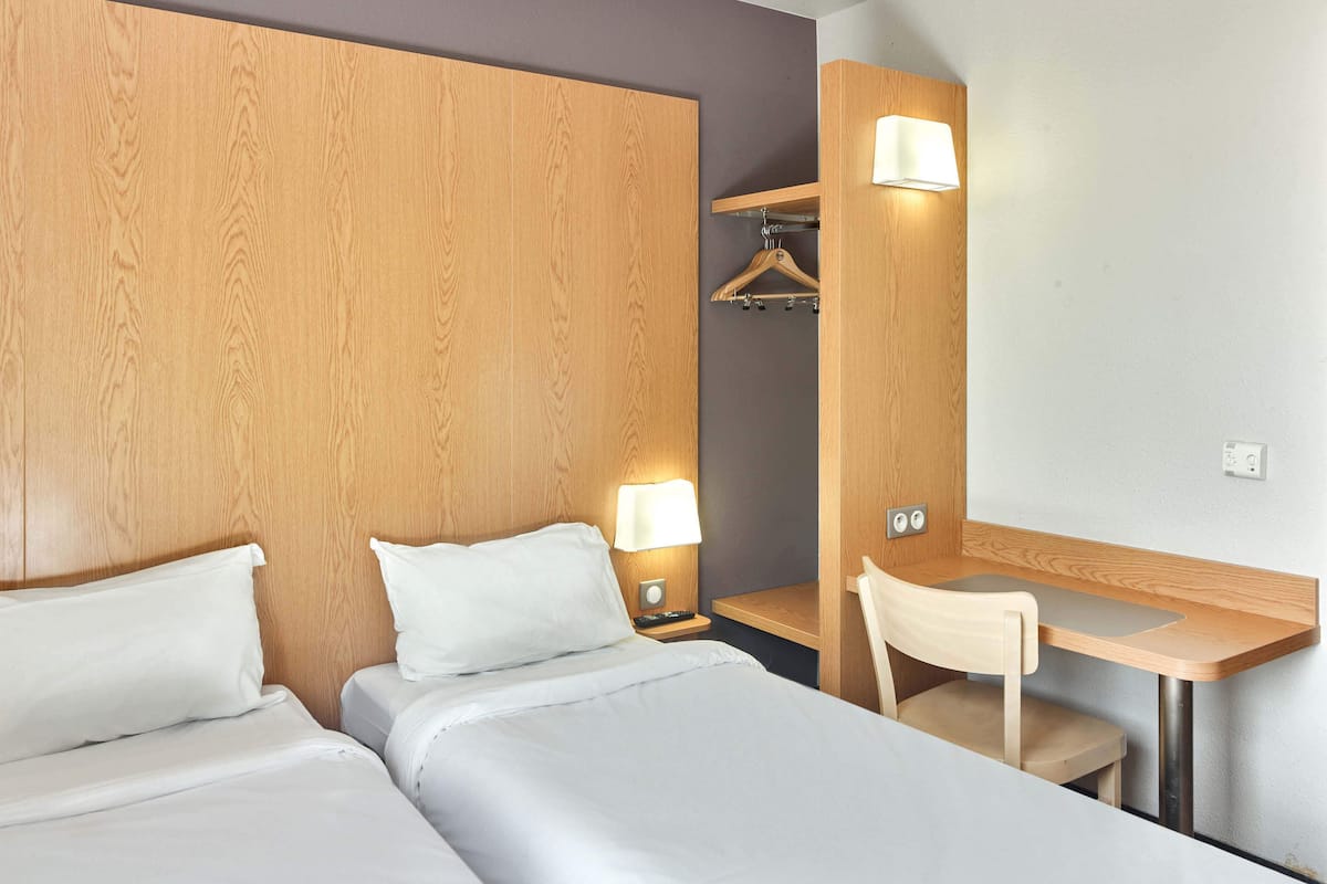 Ein Hotelzimmer mit zwei Betten, einem Holz-Kopfteil, einem Schreibtisch mit Stuhl, einem Kleiderhaken und einer Wandlampe.