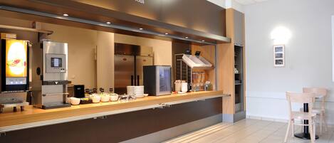 Una cafetería moderna con una estación autoservicio para café, un menú en la pizarra y un área de descanso con dos sillas de madera.