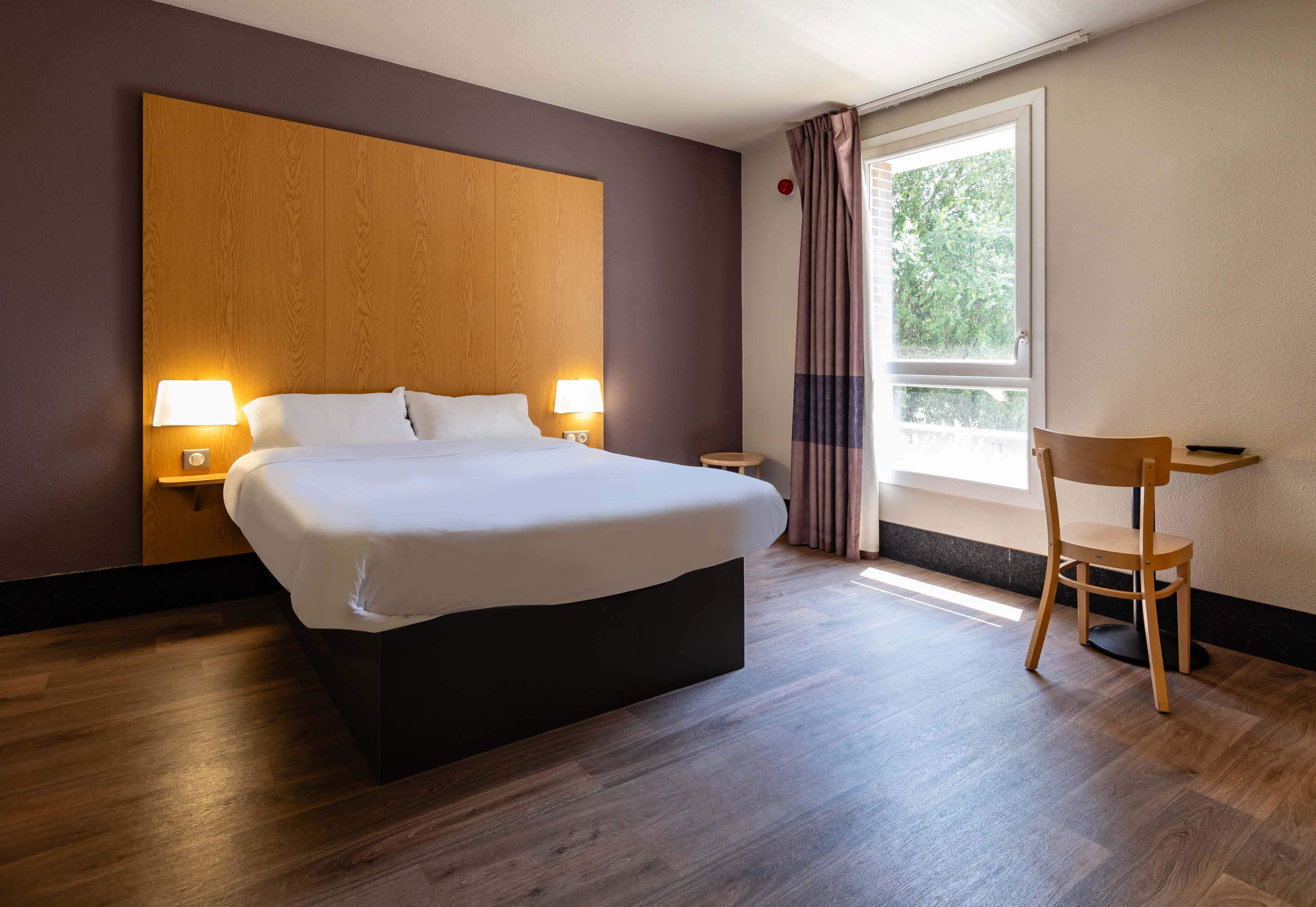 Une chambre d’hôtel moderne avec un grand lit, des lampes de chevet, une tête de lit en bois, une petite table et une chaise.