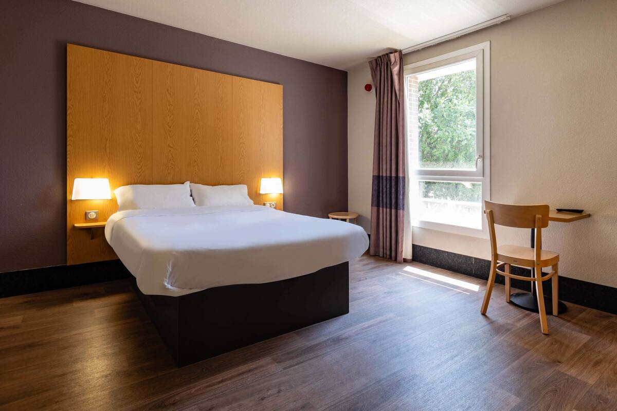 A modern hotel room with a large bed, bedside lamps, a wooden headboard, a small table, and a chair.