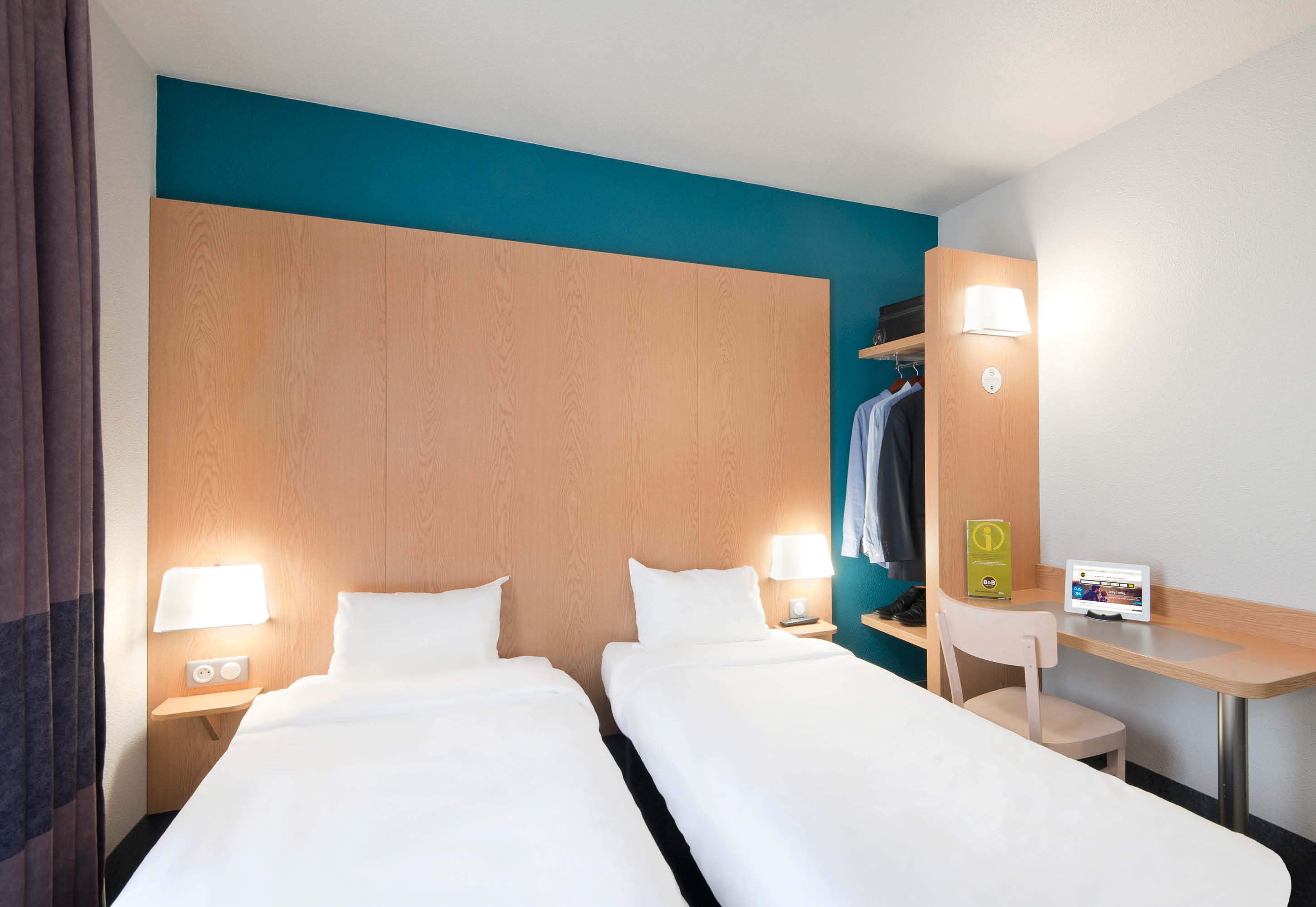 Habitación de hotel con dos camas, un escritorio, una silla, un perchero y una mesita de noche con lámpara y teléfono.