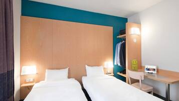 A hotel room with two beds, a desk, a chair, a coat rack, and a nightstand with a lamp and a phone.
