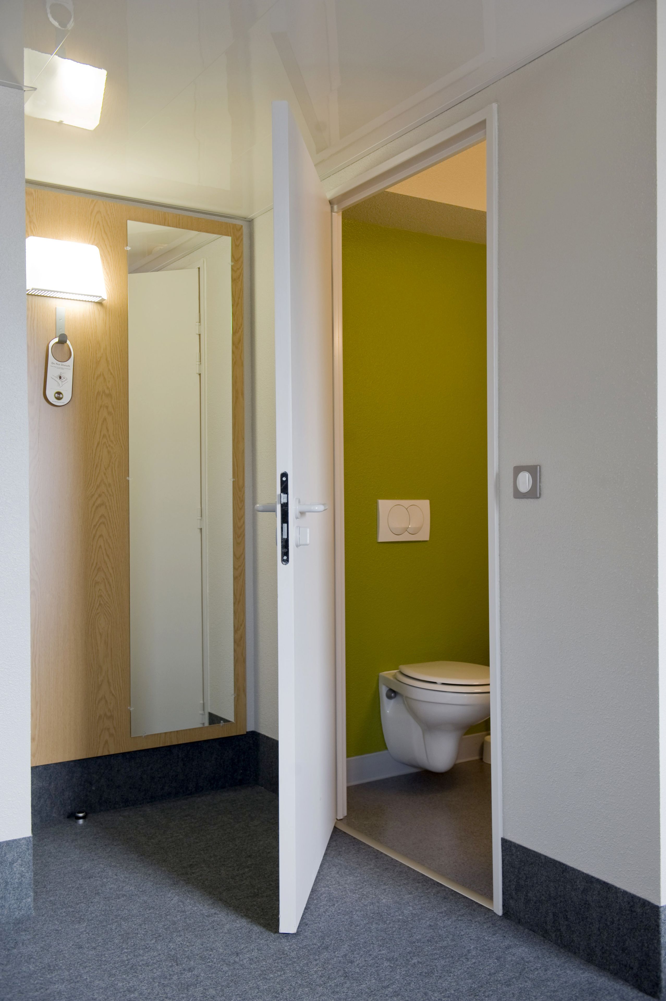 A hotel room with a wooden door, a mirror, a light fixture, a toilet, and a green wall.