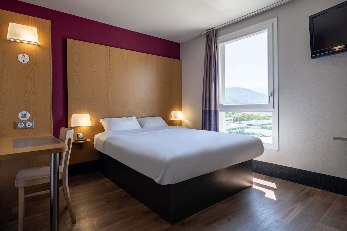 A hotel room with a bed, bedside lamps, a desk, a chair, and a window with a view.
