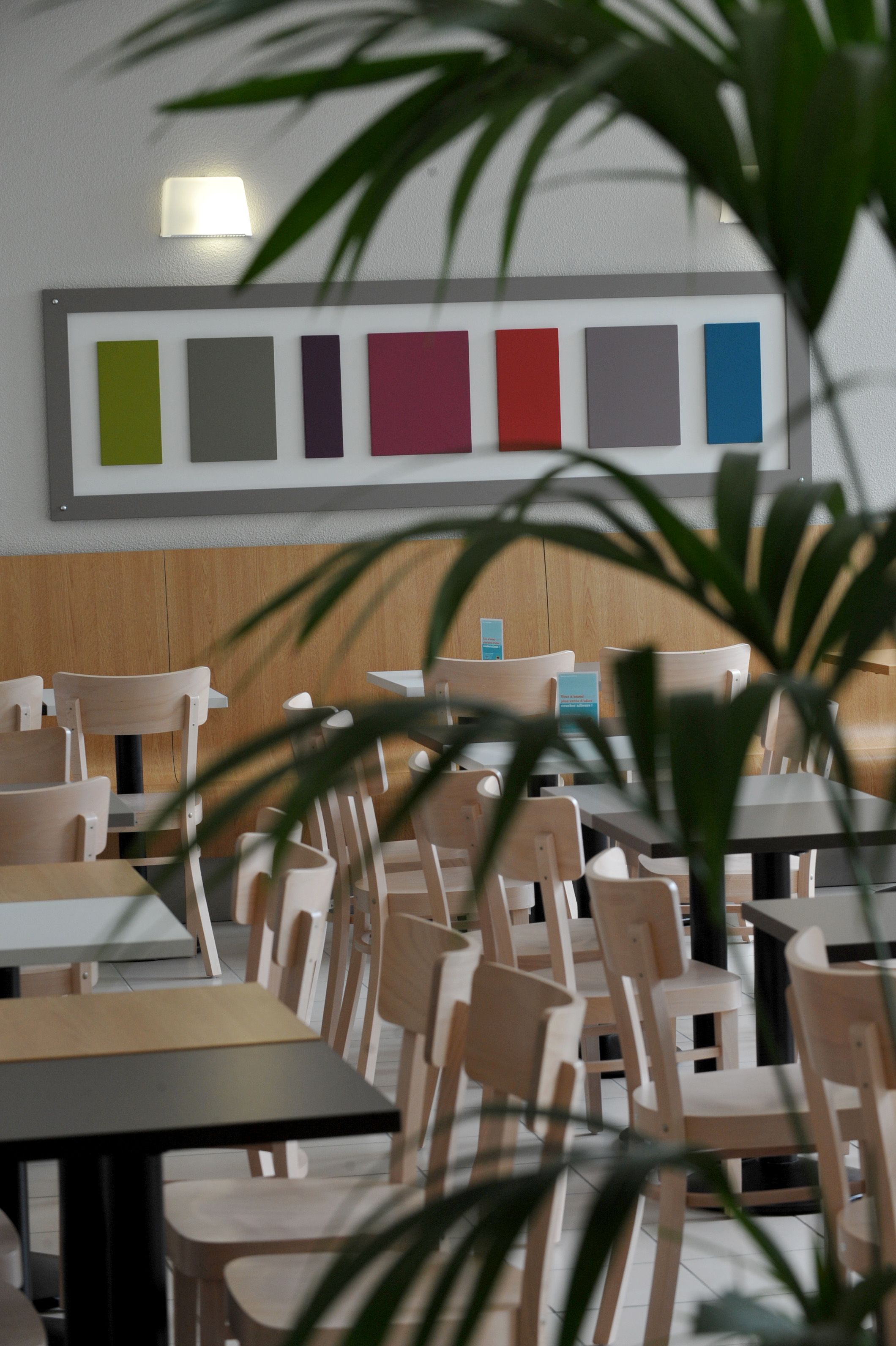 A restaurant interior with wooden chairs and tables, a wall-mounted color palette, and a potted plant in the foreground.