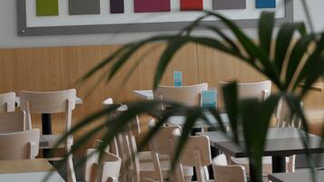 A restaurant interior with wooden chairs and tables, a wall-mounted color palette, and a potted plant in the foreground.