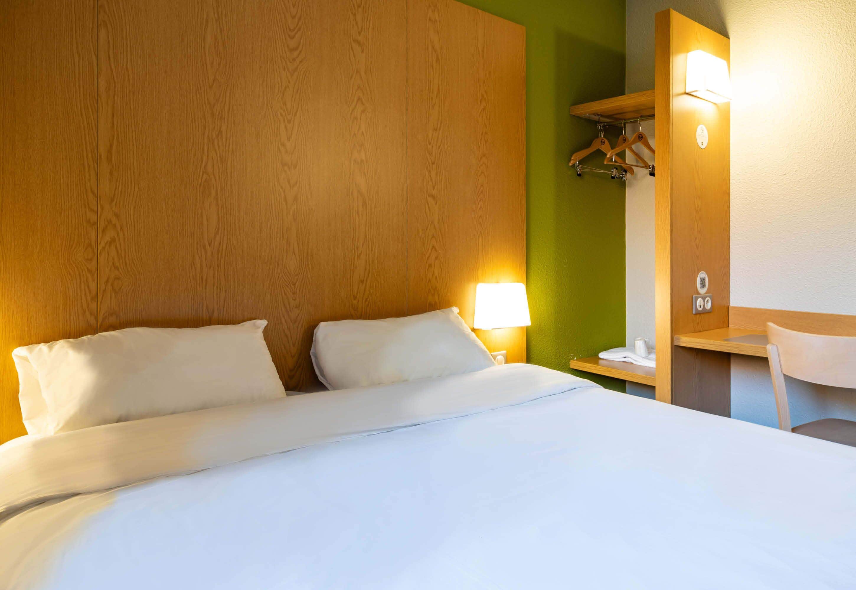 A hotel room with a wooden headboard, a bed with white linens, a bedside lamp, a desk with a chair, and a mirror.