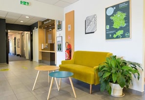 Una zona de recepció amb un sofà groc, una taula rodona petita, una planta en test i un extintor a la paret.