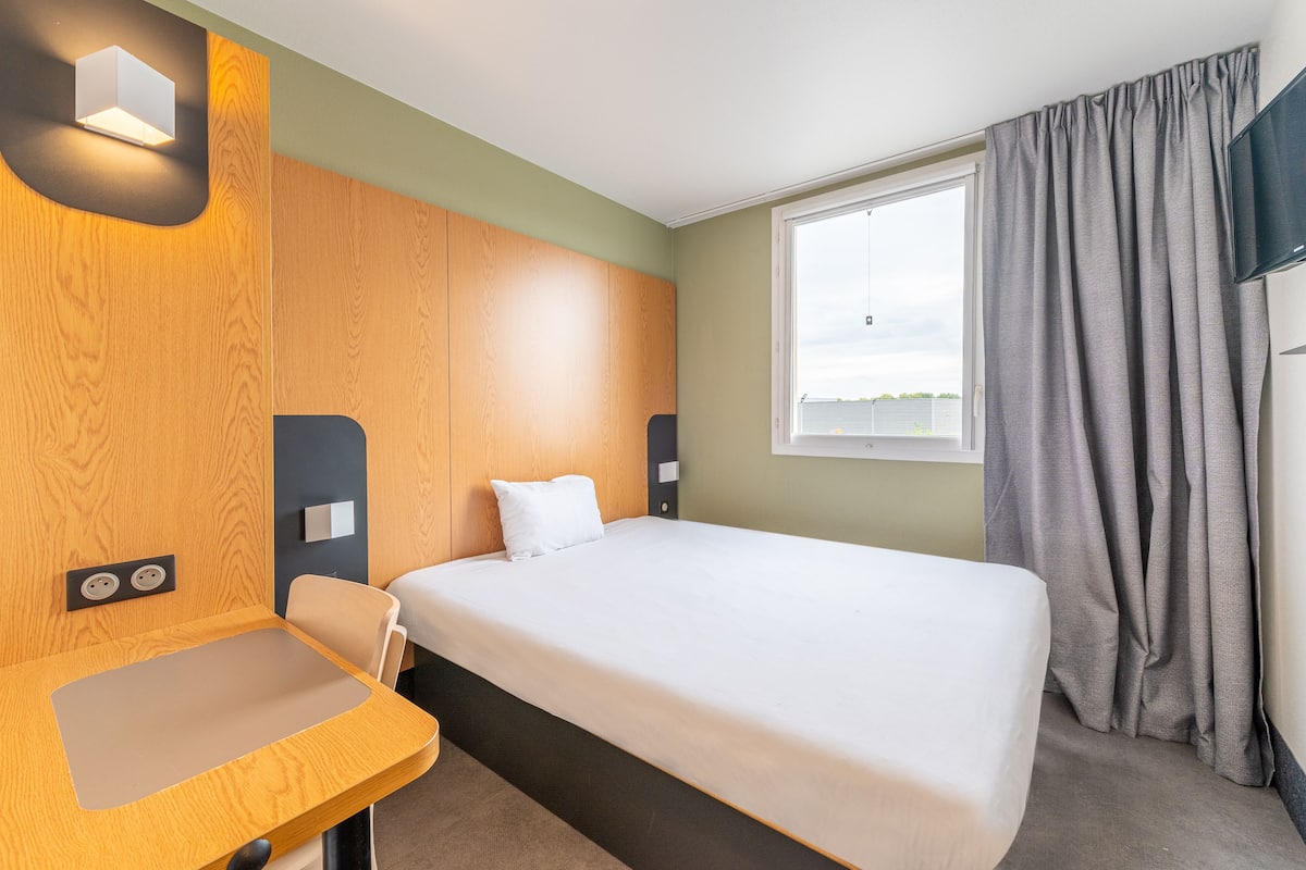 A hotel room with a bed, a desk, a chair, a window with curtains, and a TV on the wall.