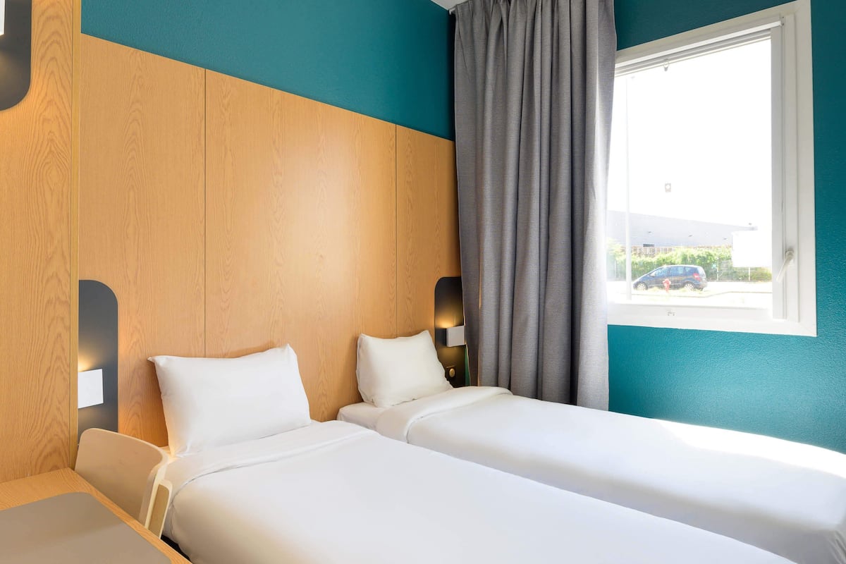 A hotel room with two beds, a window with curtains, and a view of the outside.