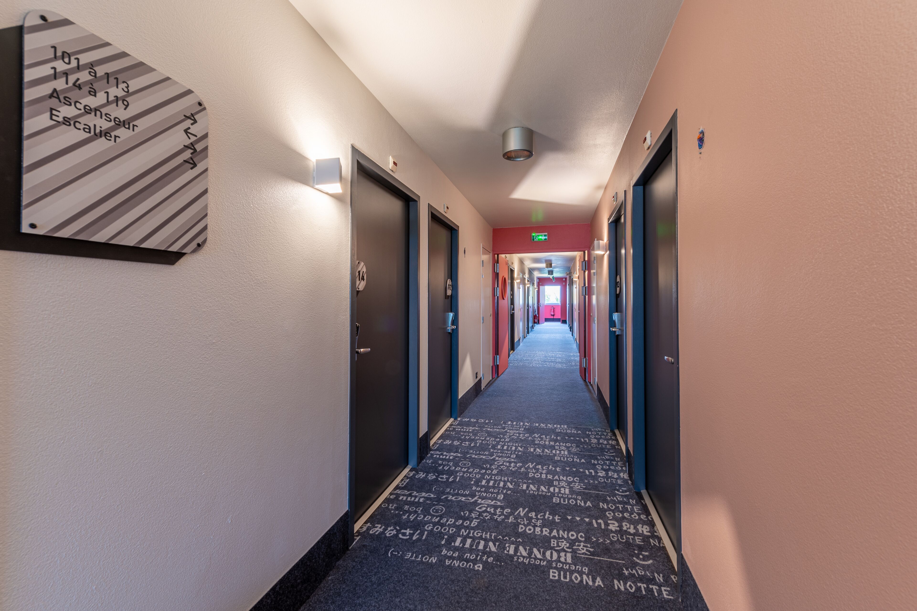 A hotel hallway with directional signs, doors, and a carpet with text.