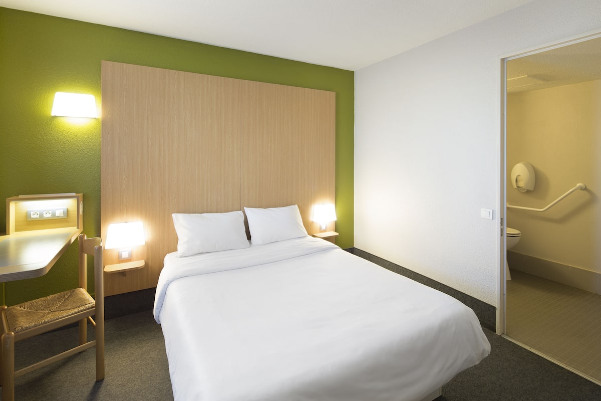 A hotel room with a bed, bedside table, lamp, chair, and a bathroom visible through an open door.