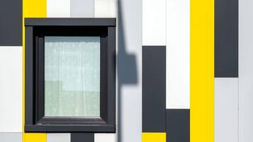 Ein Fenster mit schwarzem Rahmen und weißem Vorhang, eingelassen in eine Wand mit abwechselnd gelben und weißen Feldern.